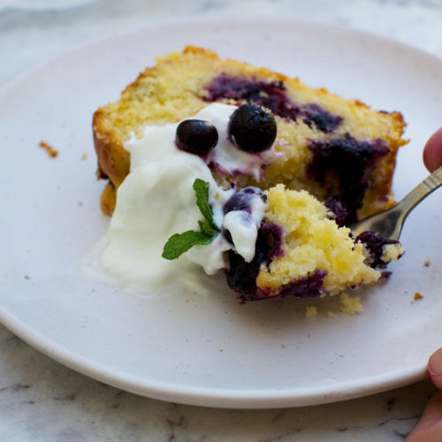 Someone eating a slice of sticky blueberry lemon curd cake with yogurt and fresh blueberries on top on a white plate with a fork.