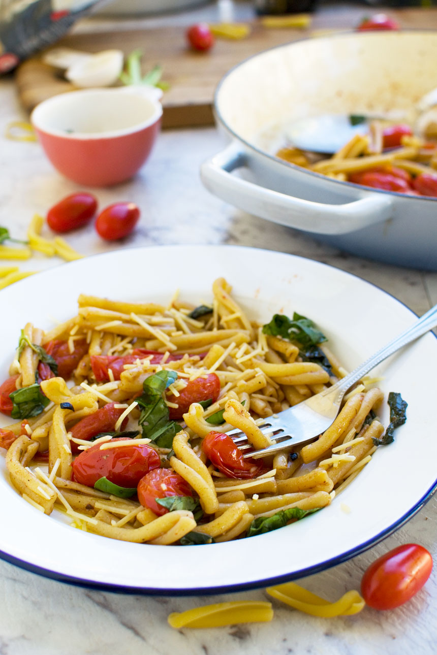 A closeup of 12-minute cherry tomato pasta with a fork in it in a white dish with a blue rim and with the edge of a baking dish in the background.