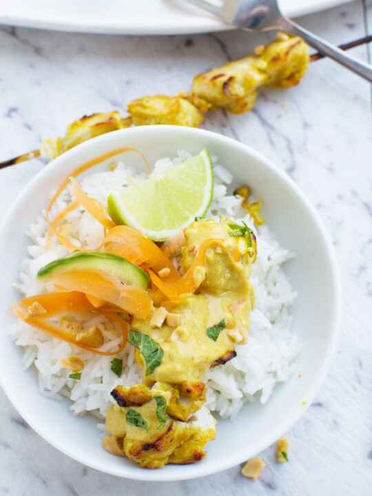 Chicken satay skewers with peanut sauce in a white bowl with rice from above on a white marble background.