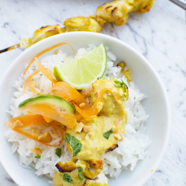 Chicken satay skewers with peanut sauce in a white bowl with rice from above on a white marble background.