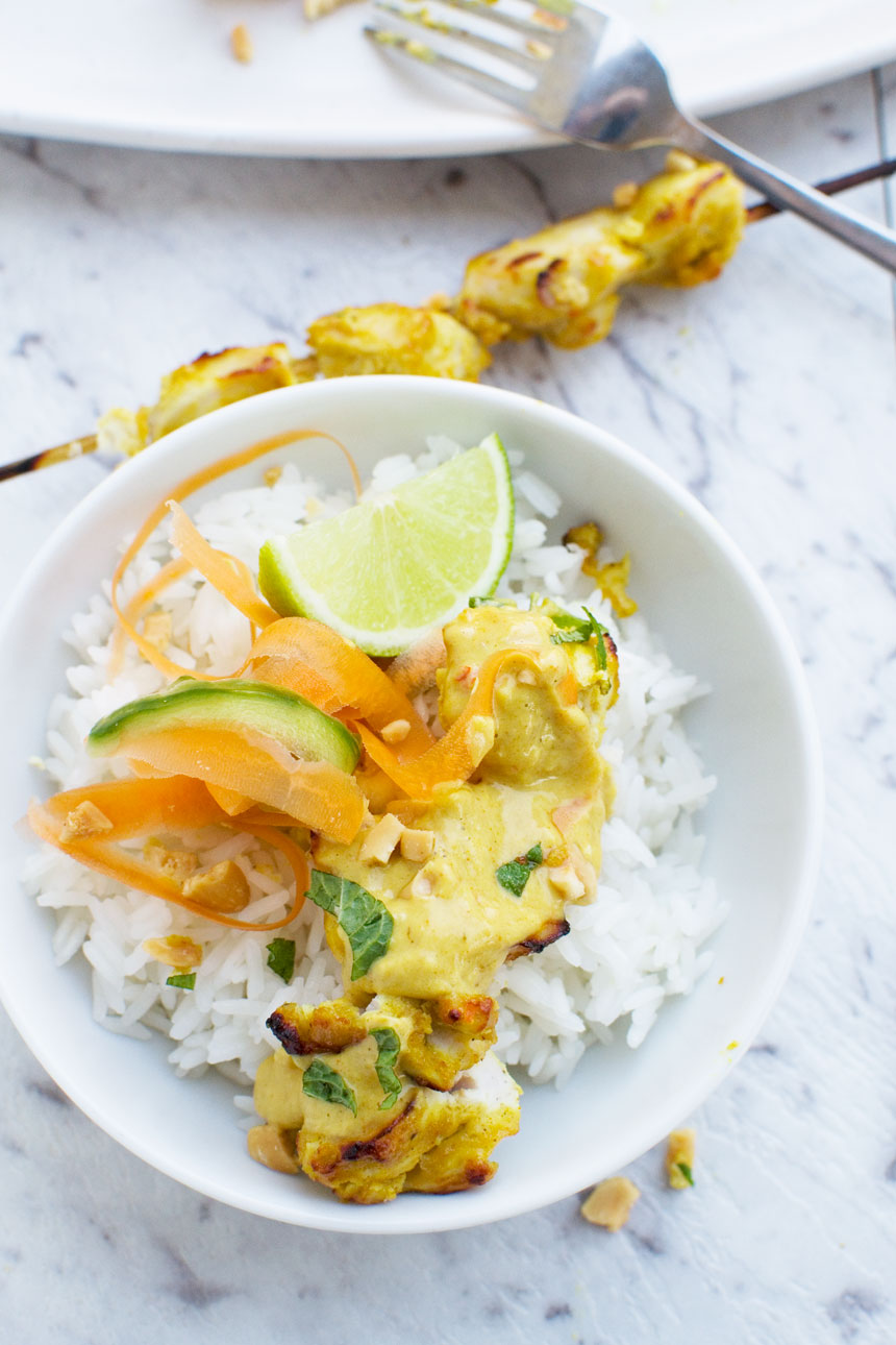 Chicken satay skewers with peanut sauce in a white bowl with rice from above on a white marble background.