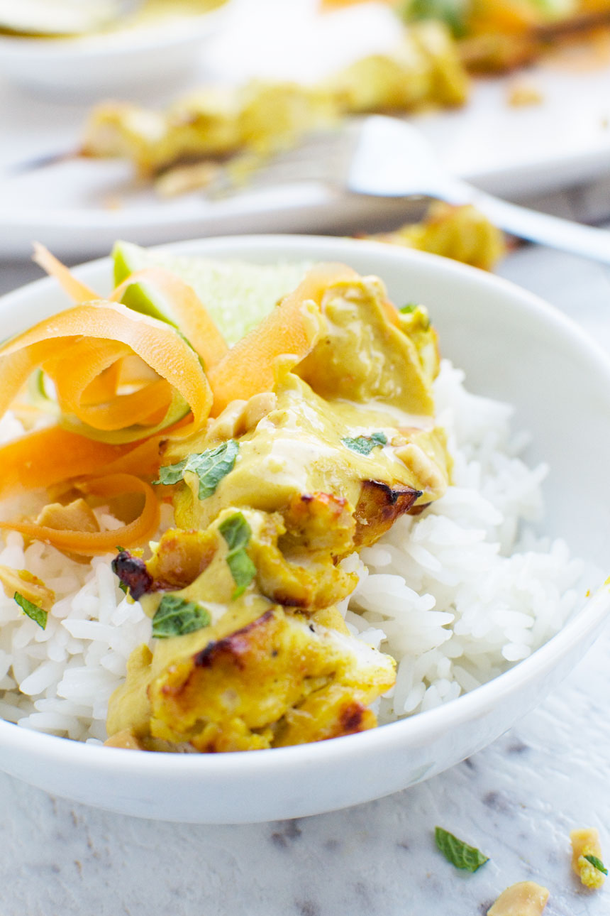 Chicken satay skewers with peanut sauce in a white bowl with rice from the front on a white marble background.