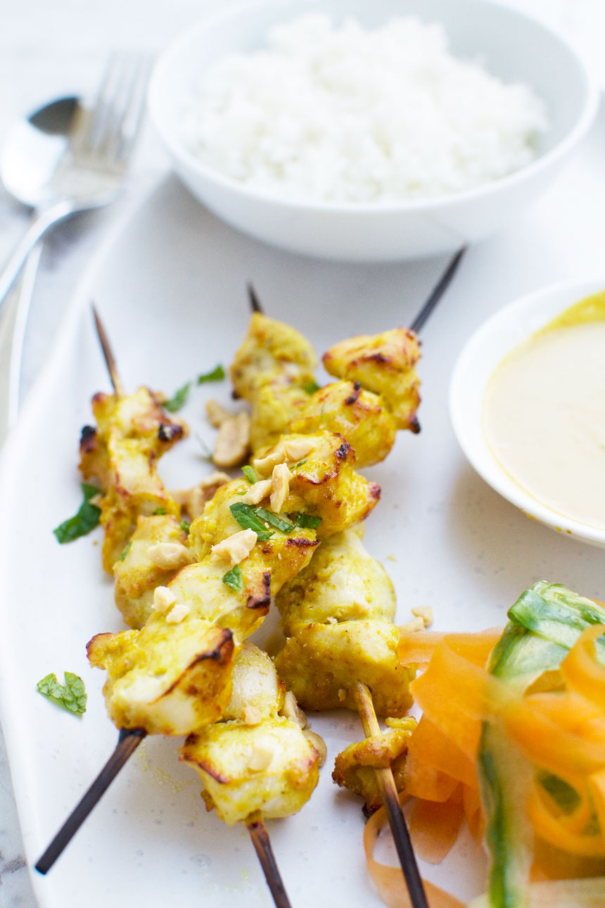 Chicken satay skewers and carrot and cucumber ribbons on a white platter with a small bowl of rice and satay sauce.