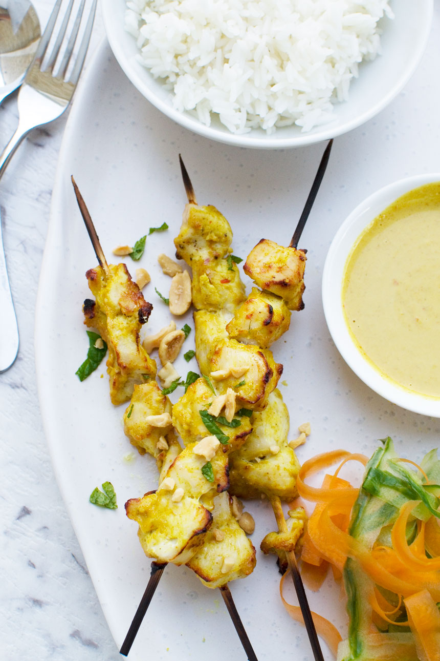 Chicken satay skewers from above on a white platter with ribbons of carrot and cucumber and with a bowl of peanut sauce on the side.