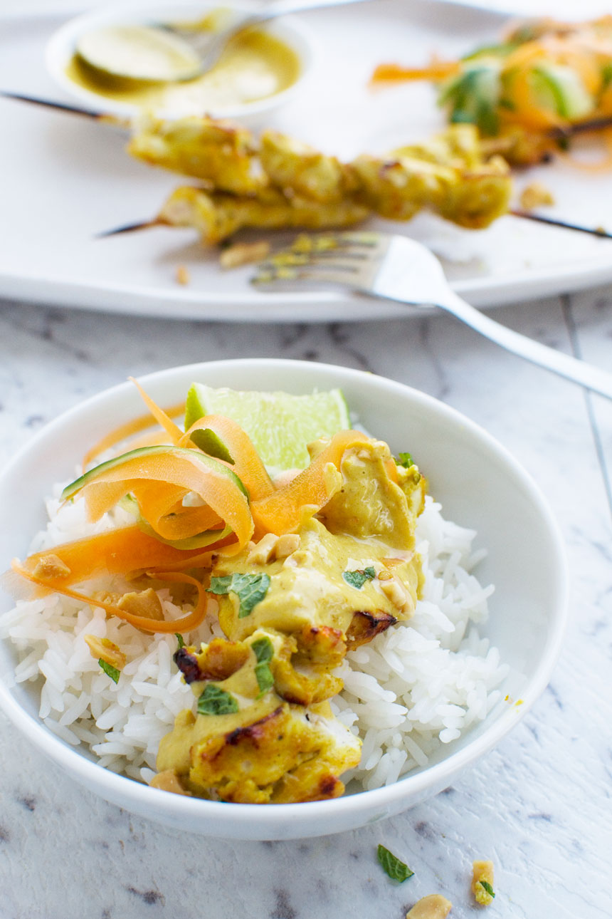 Chicken satay skewers with peanut sauce in a white bowl with rice on a white marble background with more skewers on a platter in the background.