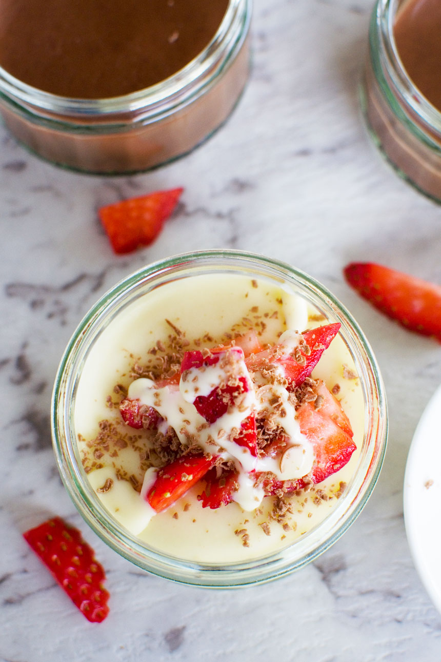 A white chocolate pot de creme with strawberries on the top on a marble background and with strawberries scattered around.