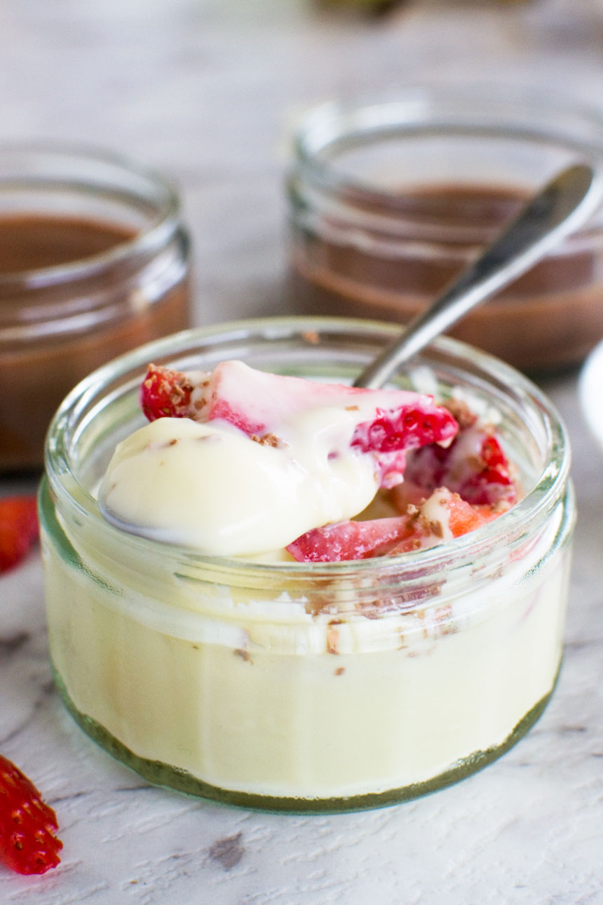 A white chocolate pot de creme with strawberries and a spoon in it with more chocolate pots in the background.