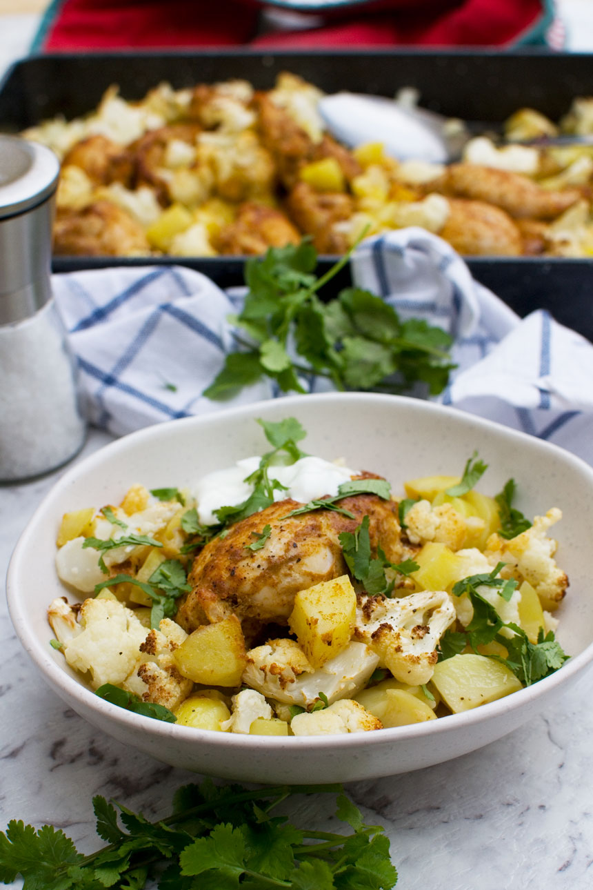 A white bowl of sheet pan chicken tikka with cauliflower and potatoes with a blue and white checked tea towel in the background and a baking tray of food.