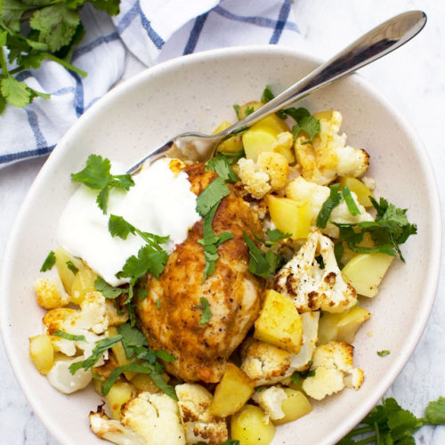 Dish of sheet pan chicken tikka with cauliflower and potatoes from above in a white dish with a spoon and yogurt in it and a blue white checked tea towel.