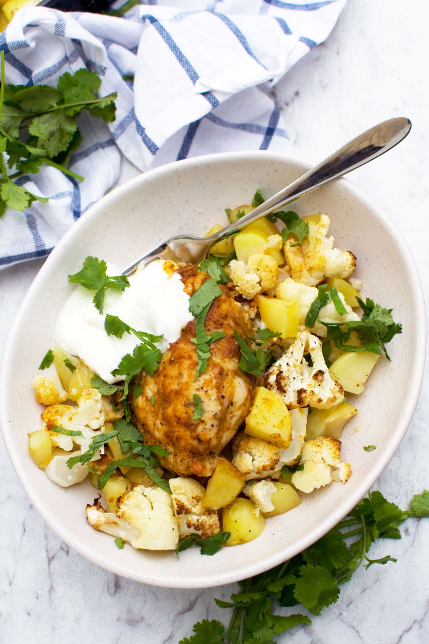 Dish of sheet pan chicken tikka with cauliflower and potatoes from above in a white dish with a spoon and yogurt in it and a blue white checked tea towel.