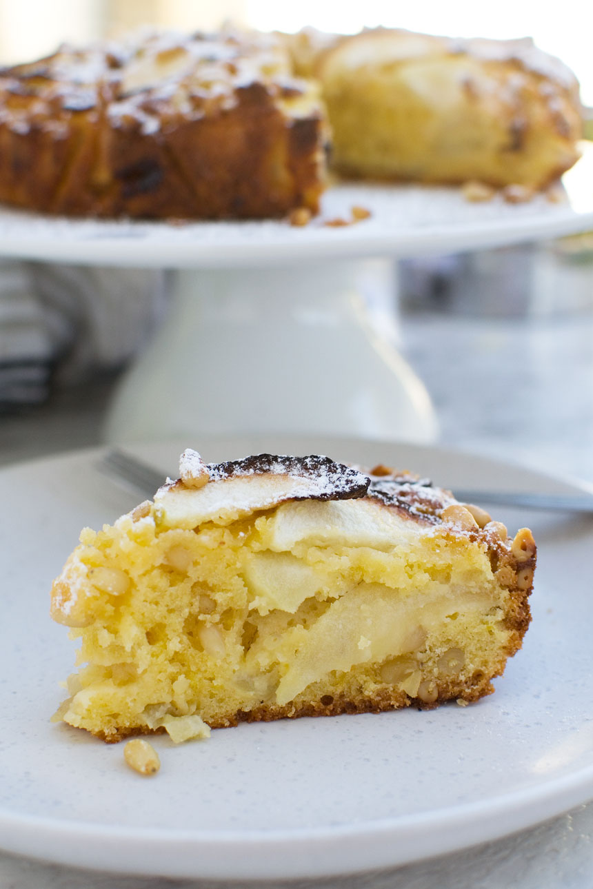a piece of easy apple cake (torta di mele) on a  white plate with more cake in the background on a white cake stand