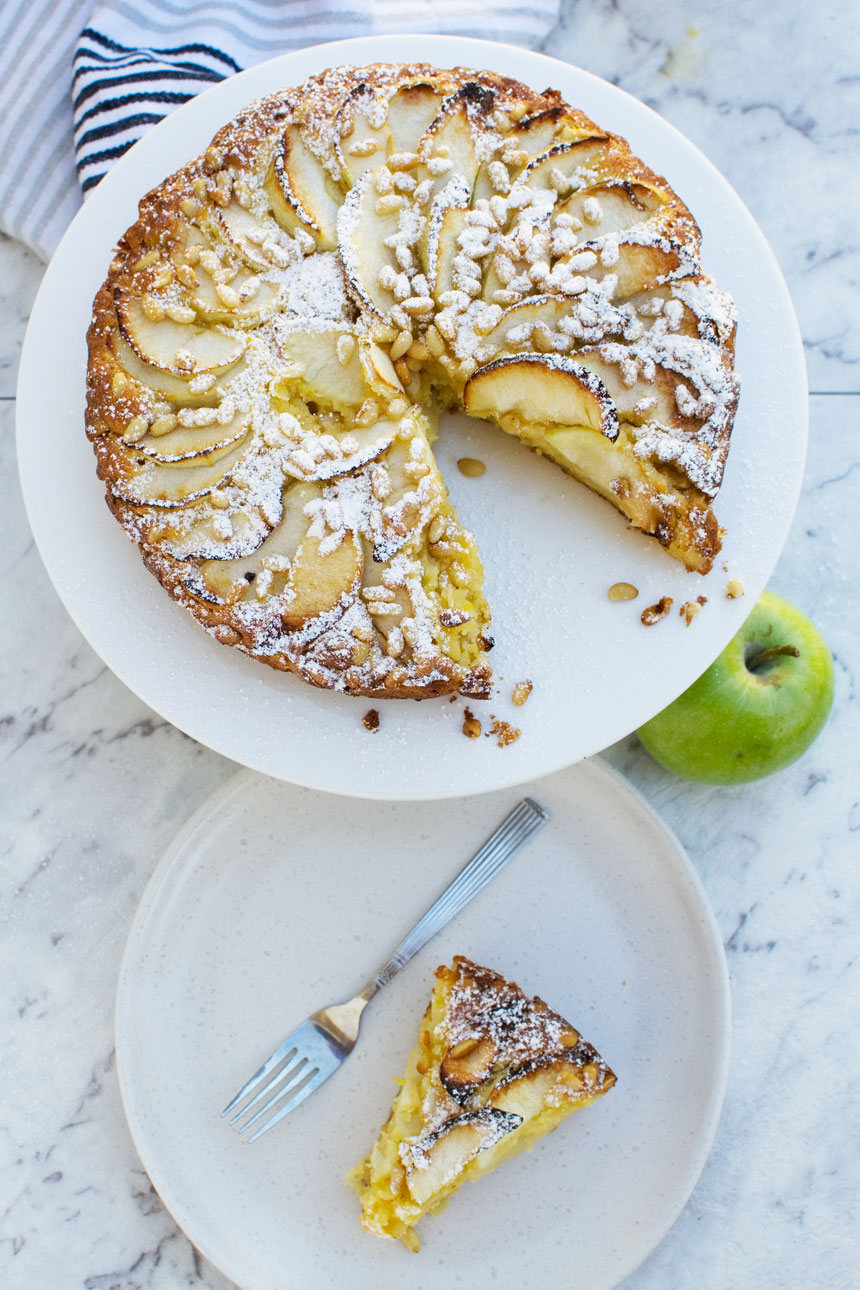 Easy apple cake (torta di mele) on a white plate and marble background with an apple and a single piece on a plate from above