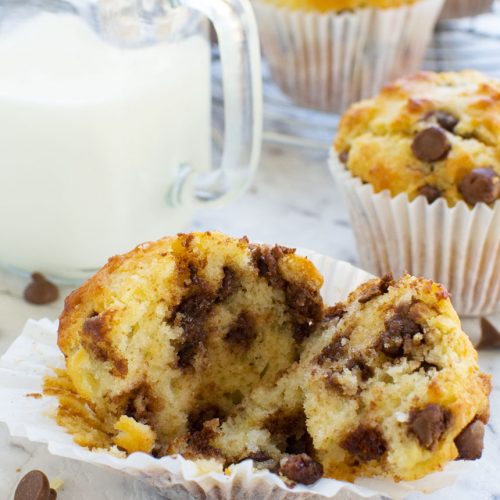 A Greek yogurt choc chip muffin split open in its case with milk and more muffins in the background