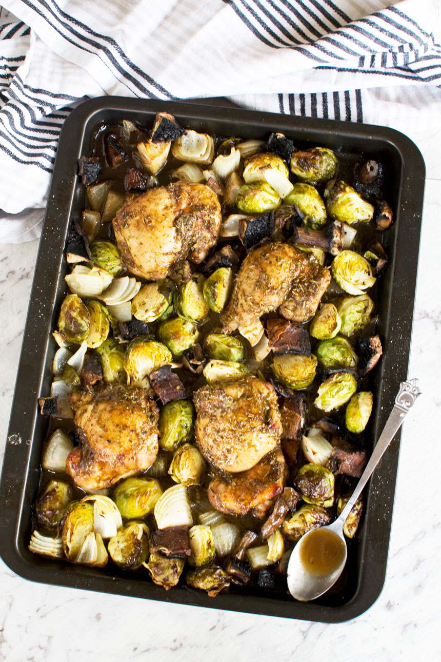 A baking tray of Honey balsamic chicken with mushrooms and brussels sprouts from above with a striped tea towel next to it