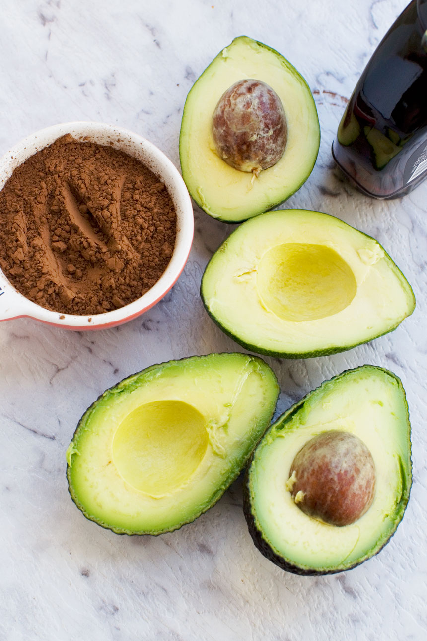 ingredients for Healthy chocolate frosting including sliced open avocados, cocoa and maple syrup on a white marble background