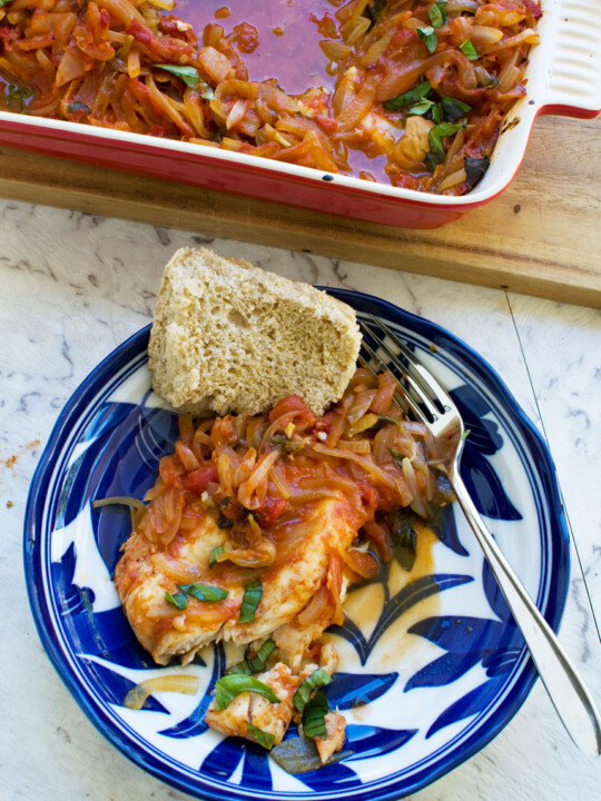 Greek baked fish in a patterned blue dish with a piece of bread and the red baking dish in the corner of the image from above