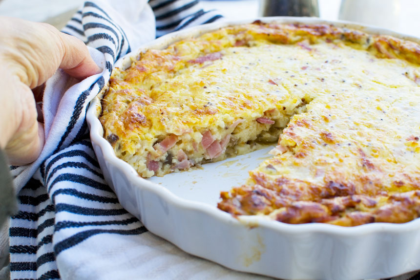 A whole crustless quiche (impossible quiche) with a piece out of it in a round baking dish with a blue striped tea towel