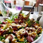 A person serving warm butternut squash, chorizo and feta salad with salad servers