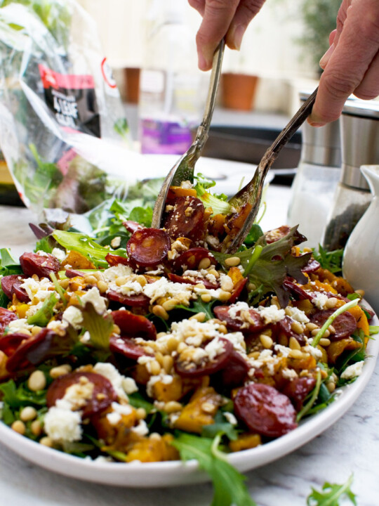 A person serving warm butternut squash, chorizo and feta salad with salad servers