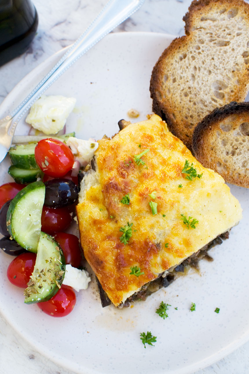 An overhead shot of a piece of easy moussaka on a white plate with bread and Greek salad 