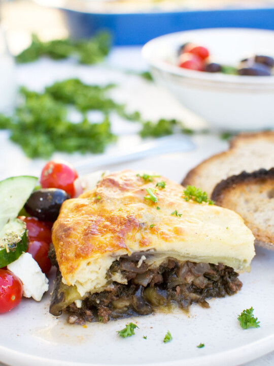 A piece of easy moussaka on a white plate with bread and Greek salad