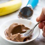 Someone eating 5-minute chocolate banana ice cream with a spoon with a banana and ice cream scoop in the background