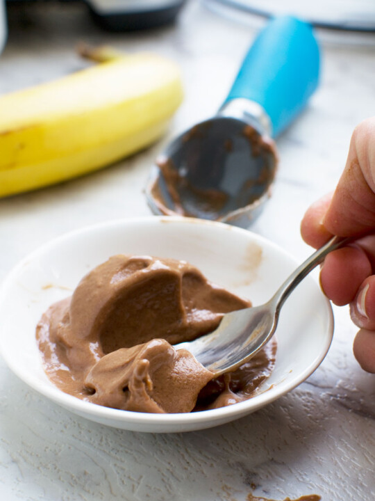 Someone eating 5-minute chocolate banana ice cream with a spoon with a banana and ice cream scoop in the background