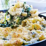 Someone serving a big spoon of Greek mac and cheese bake out of a black cast iron pan