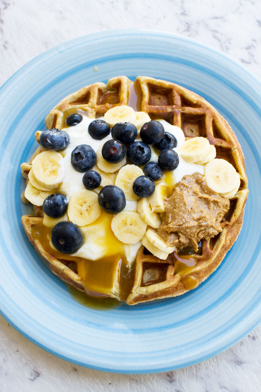 An overhead photo of a round waffle topped with yogurt, bananas, blueberries and peanut butter