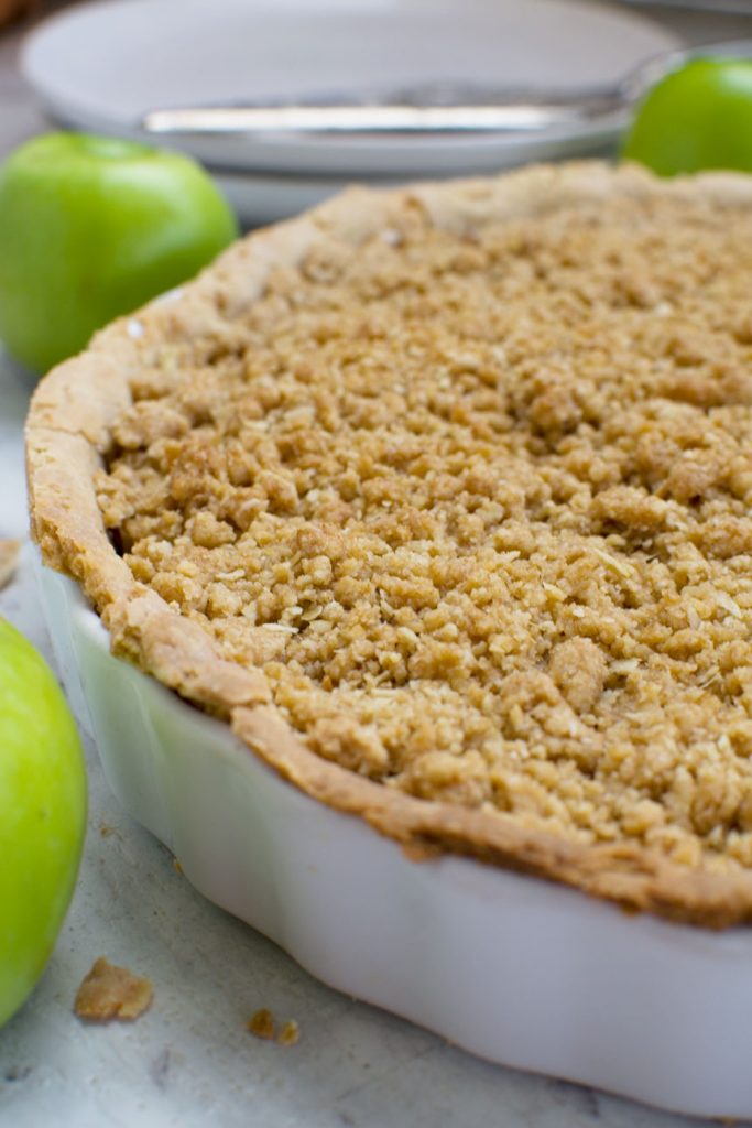 A close of of a whole easy apple crumble pie in a pie dish with apples in the background
