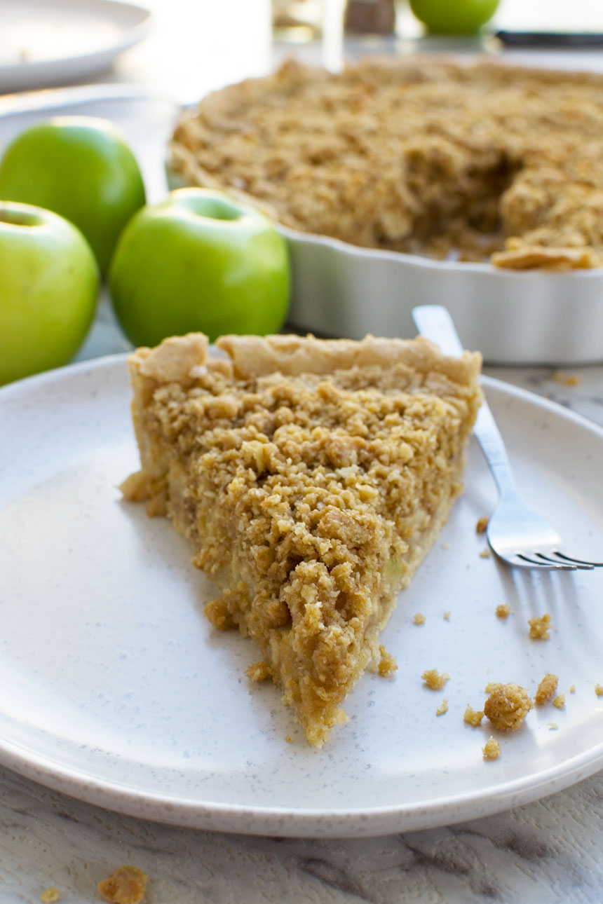 A close up of a piece of apple crumble pie on a white plate with a fork and with green apples in the background and the rest of the pie