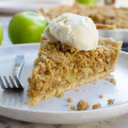A piece of easy apple crumble pie on a white plate with ice cream on top and apples in the background
