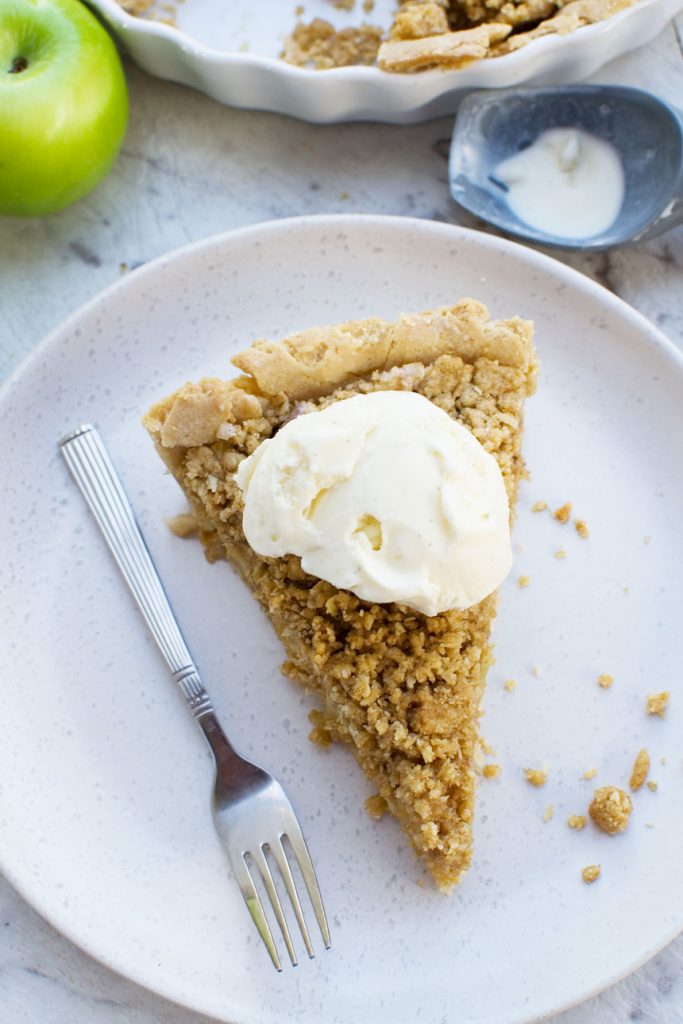 A close up of a piece of apple crumble pie on a white plate with ice cream on top - overhead