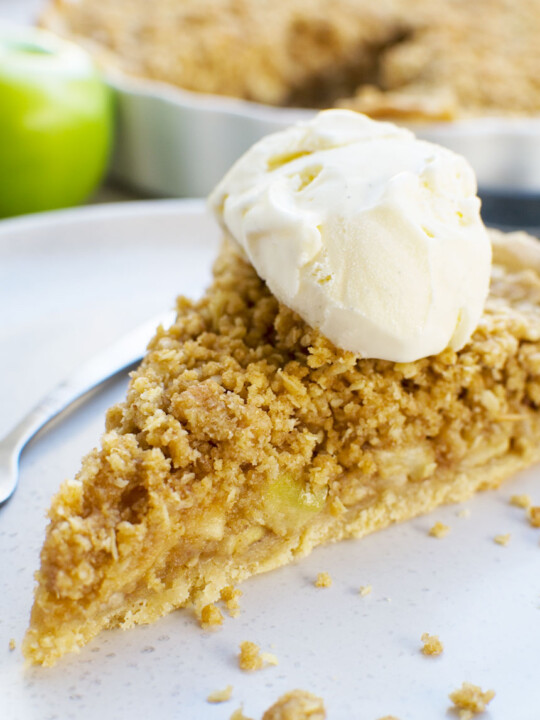 A close up of a piece of easy apple crumble pie on a white plate with ice cream on top and apples in the background