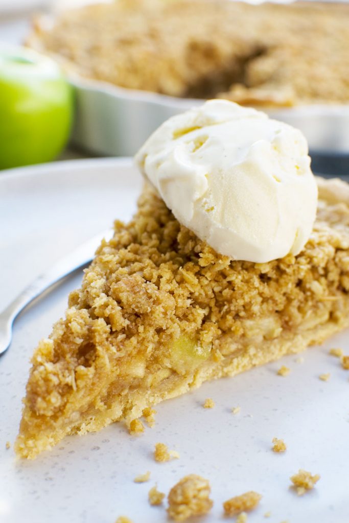 A close up of a piece of easy apple crumble pie on a white plate with ice cream on top and apples in the background