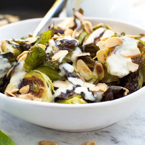 A white bowl filled with crispy brussels sprouts with tahini sauce and almonds