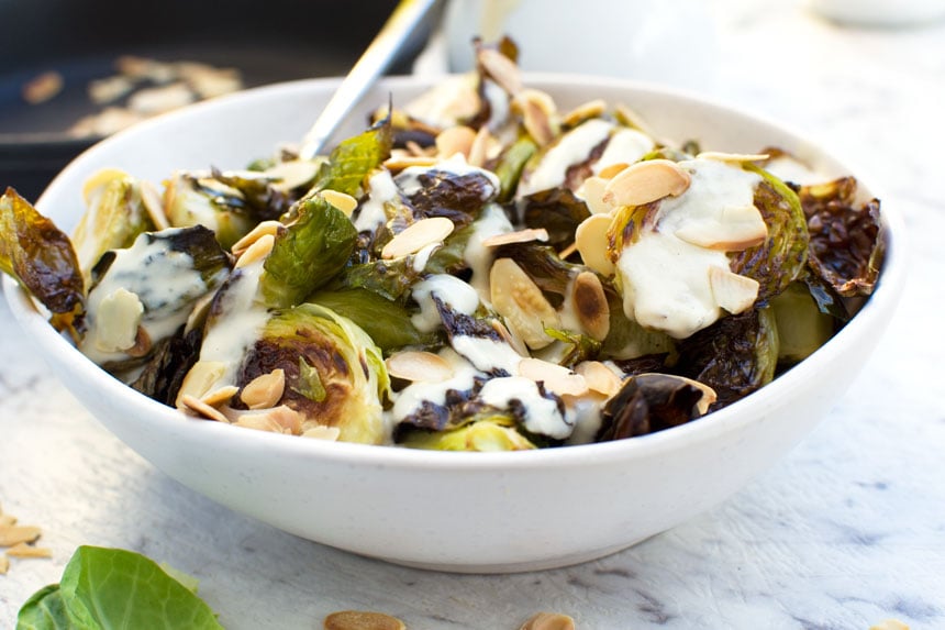 A white bowl filled with crispy brussels sprouts with tahini sauce and almonds