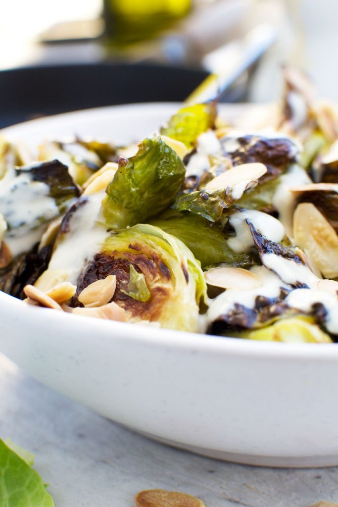 A close up of crispy brussels sprouts with tahini sauce and almonds in a white bowl