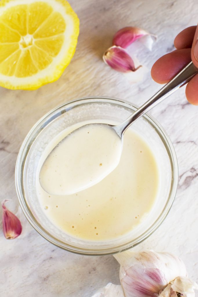 A small bowl of tahini dressing with a spoon being held over it and a lemon in the background