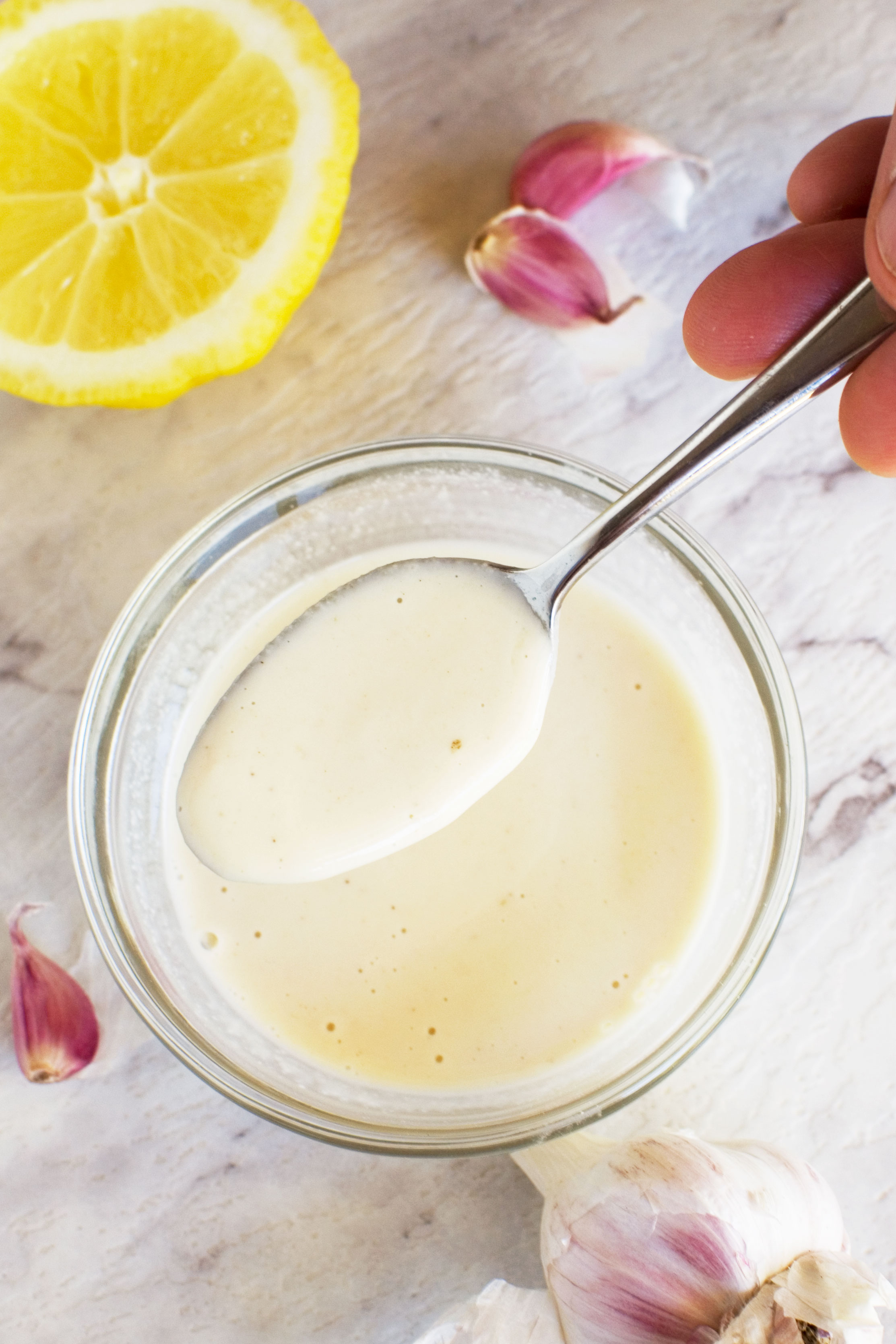 A small bowl of tahini dressing with a spoon being held over it and a lemon in the background