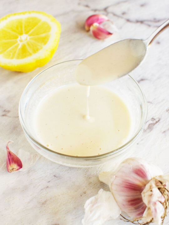 A spoon drizzling tahini dressing into a small bowl of it with lemon and garlic cloves in the background