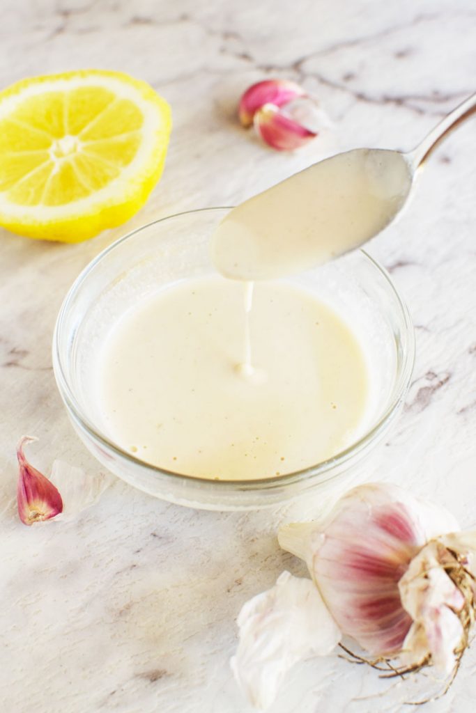 A spoon drizzling tahini dressing into a small bowl of it with lemon and garlic cloves in the background