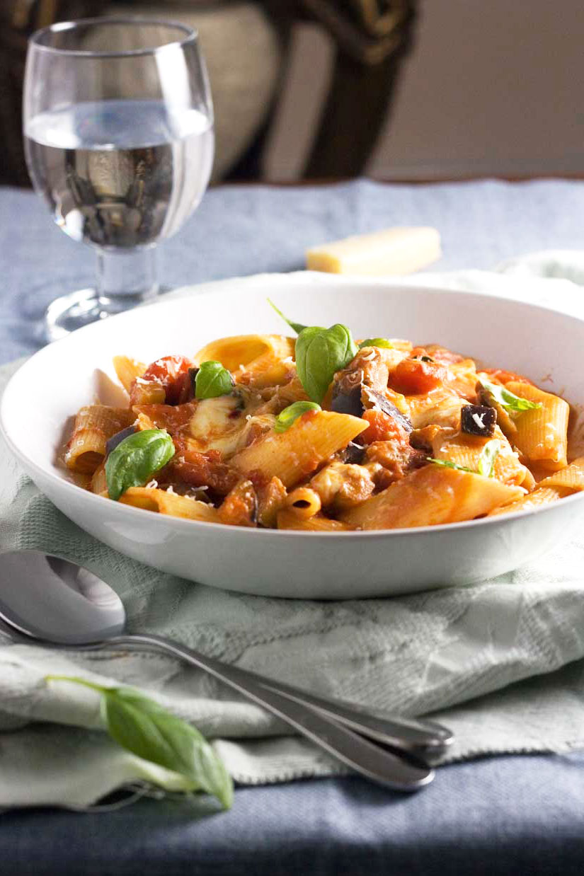 A plate of Pasta alla Norma on a light green cloth with a glass in the background