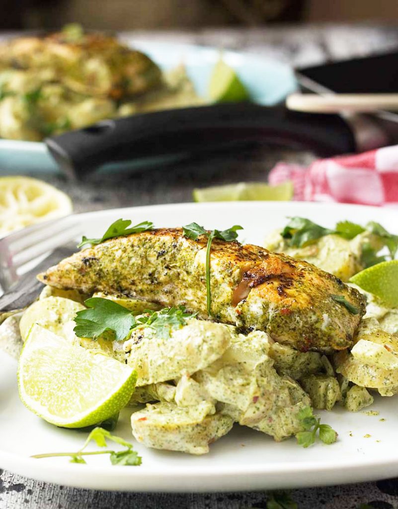 jerk salmon with yoghurt potatoes and a lime wedge on a white plate with a fork and with equipment and more food in the background.