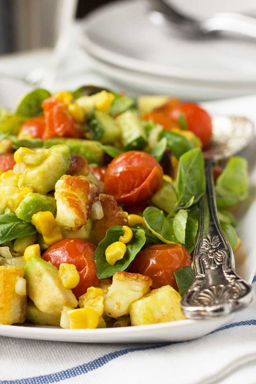 A crispy halloumi salad with avocado, corn and tomatoes on a white rectangular plate and a fancy serving spoon