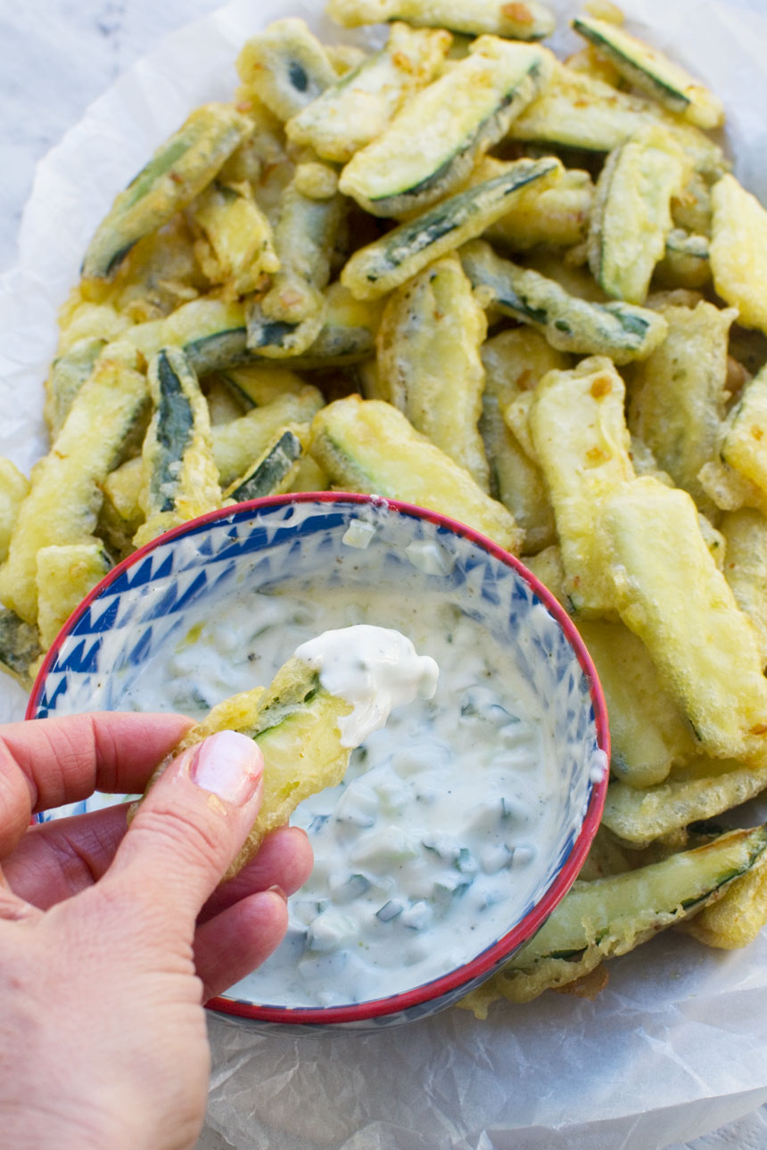 A person dipping a zucchini fry from a big plate into a bowl of tzatziki sauce