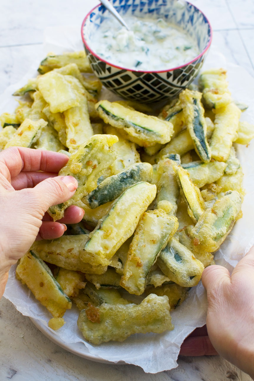 A hand dipping a zucchini fry from a big plate of them into tzatziki sauce