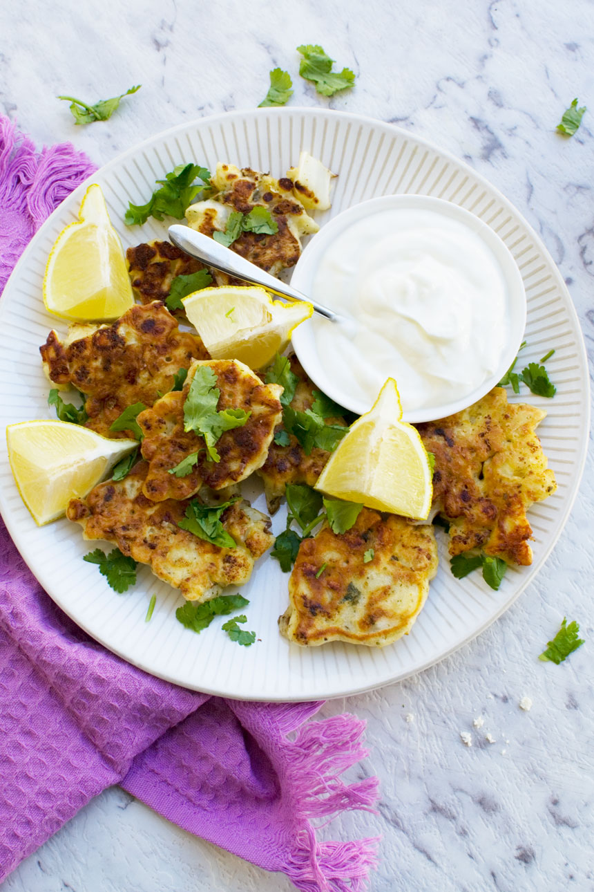 A white plate of cauliflower fritters with feta and mint on a marble background with a purple tea towel