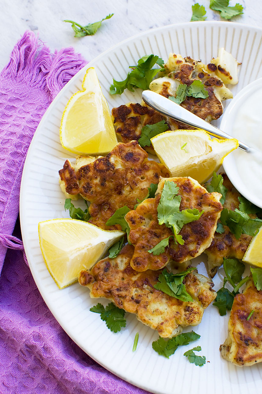 A closeup of a white plate of cauliflower fritters with lemon with a purple tea towel in the background