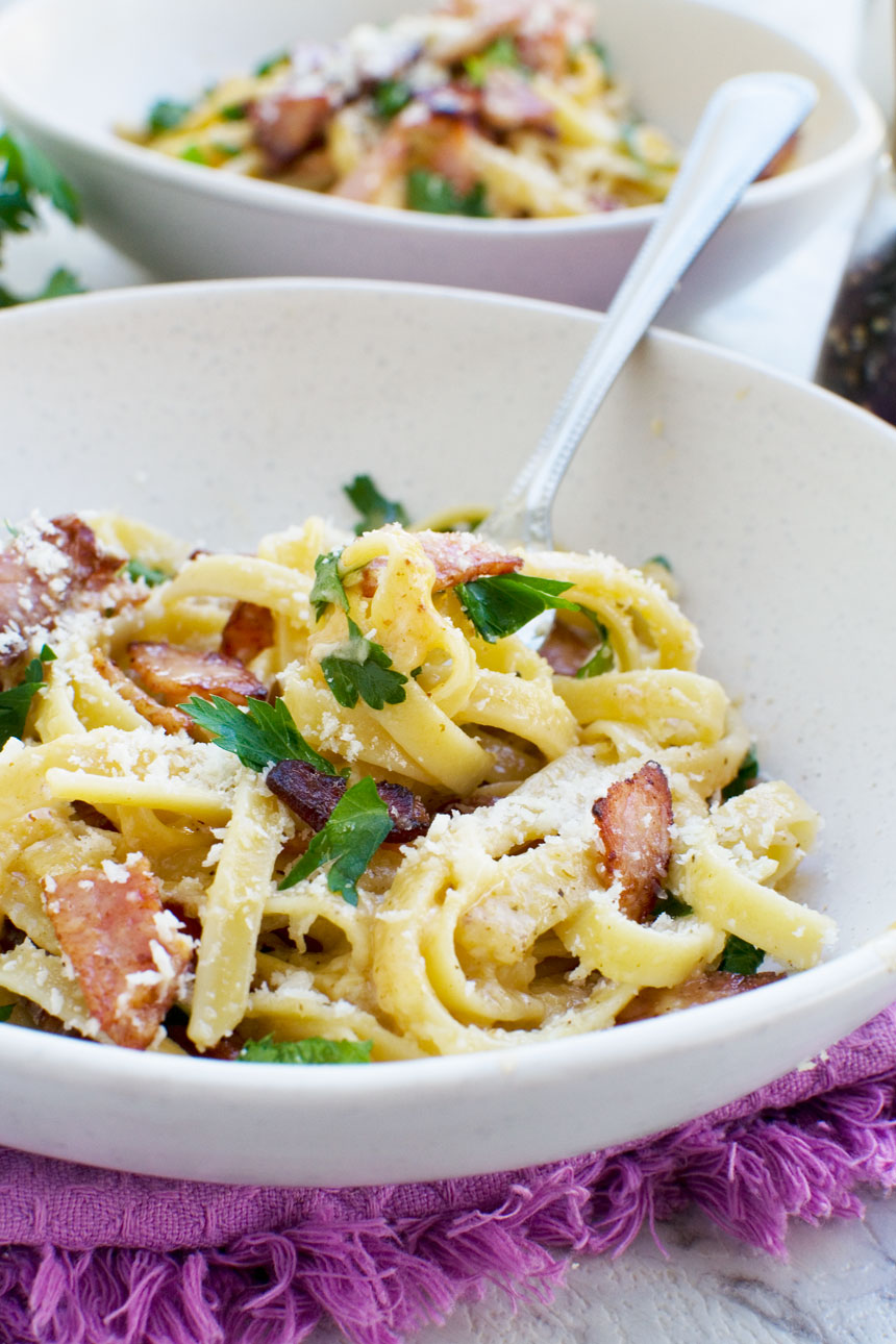 A closeup of a fork in a bowl of pasta carbonara with another bowl in the background